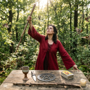 Frau in rotem Gewand steht mit erhobenem Stab hinter einem Holztisch im Wald auf dem Kelch, Dolch, Pentagramm, Tarotkarten und Kerze liegen (KI-generiert)